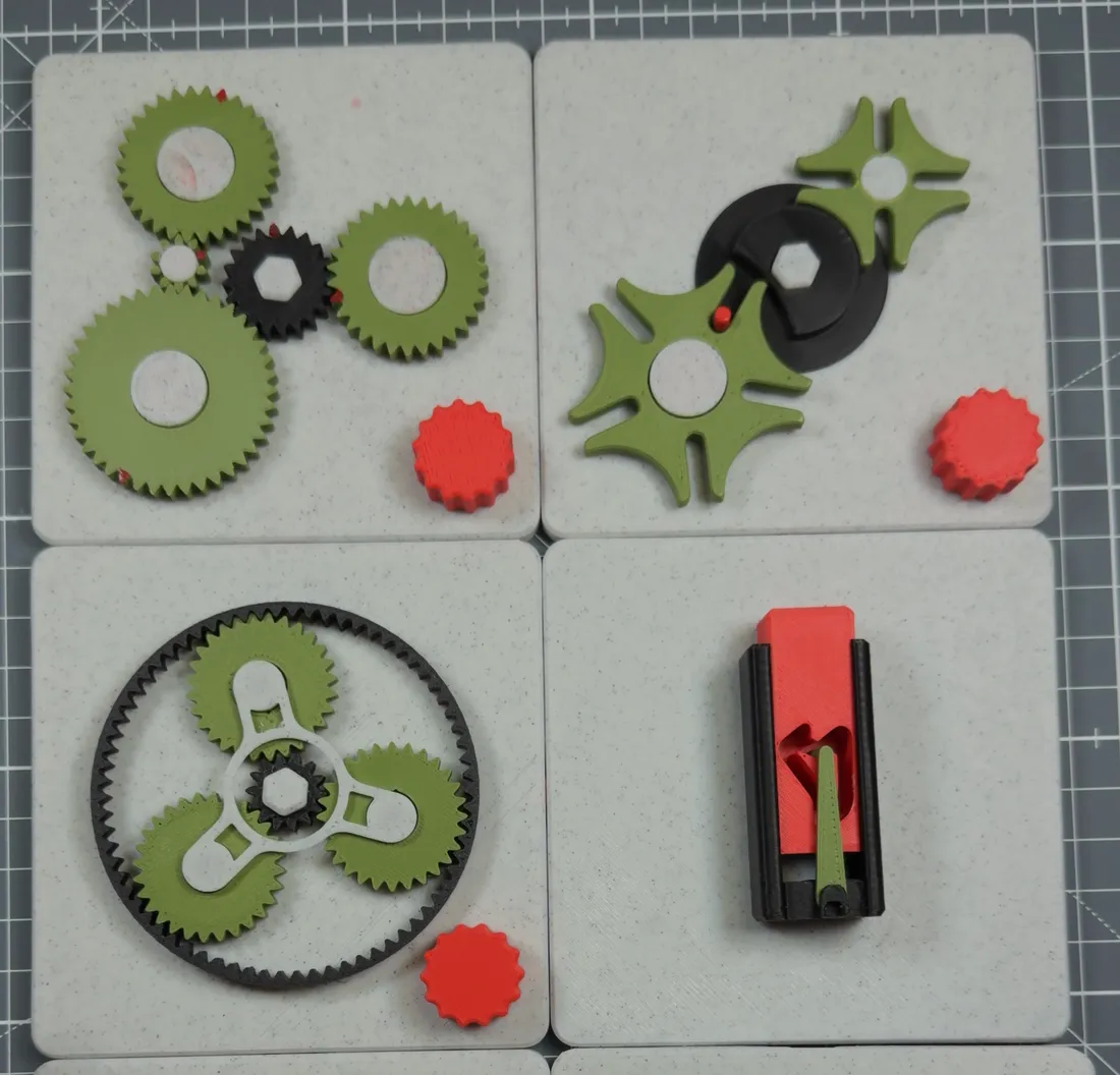Four tiles clicked together on a cutting mat: basic gears, a Geneva mechanism, a planetary gear set and a slotted latch.